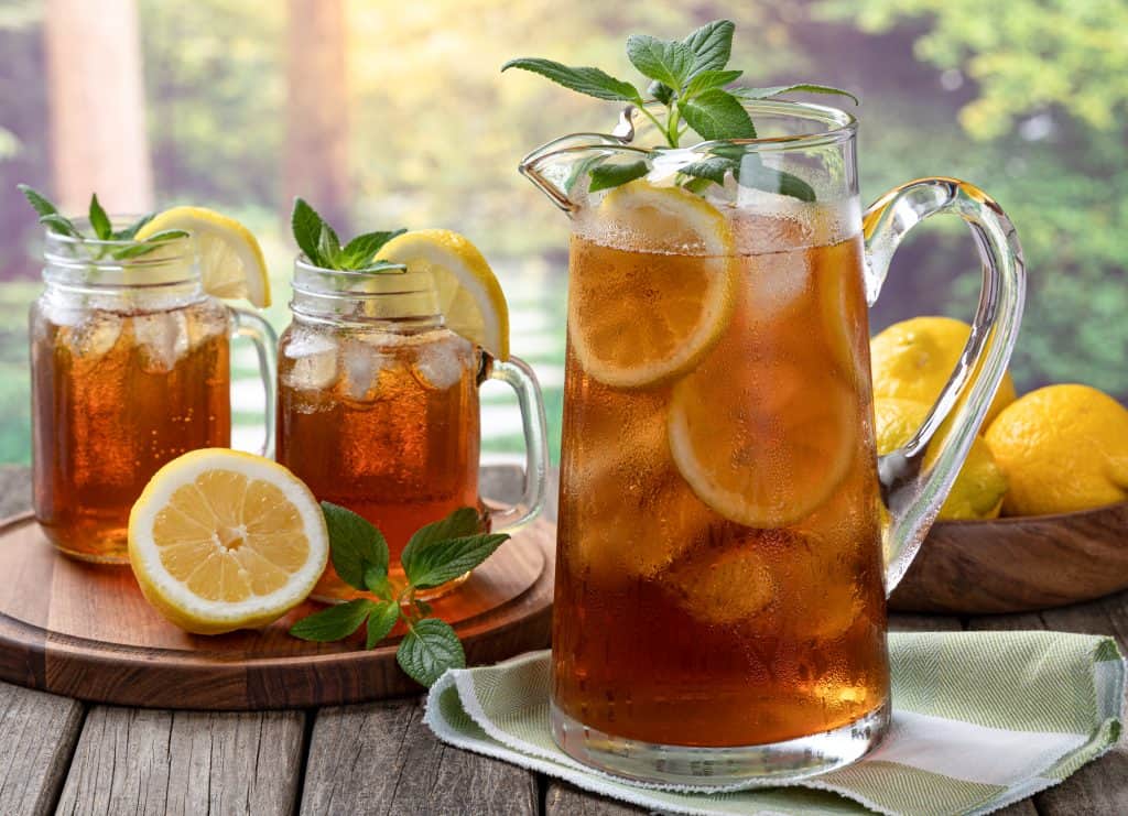 Pitcher of cold iced tea with mint, lemon slices and ice with two glasses ot tea on a wooden table and rural summer background
