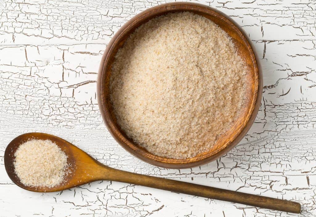 Heap of psyllium husk also called isabgol in wooden bowl and spoon on white table background. Psyllium husk also called isabgol is fiber derived from the seeds of Plantago ovata plant found in India. Flat lay from above.