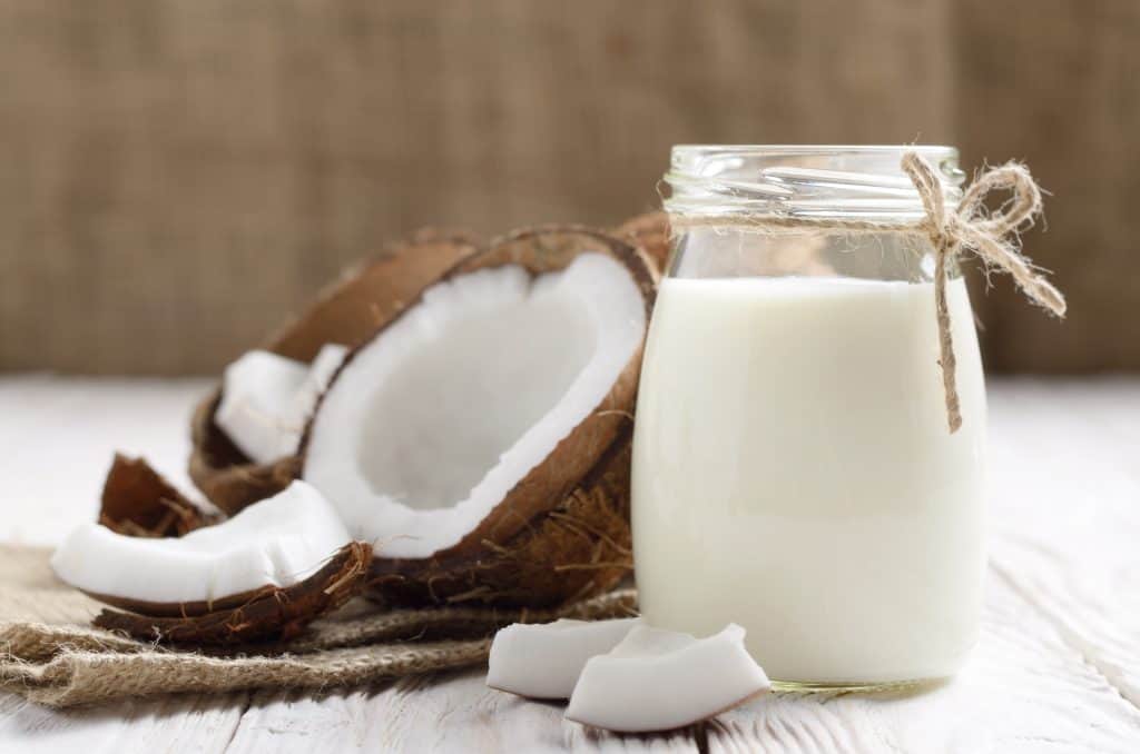 Mason jar of milk or yogurt on hemp napkin on white wooden table with coconut aside