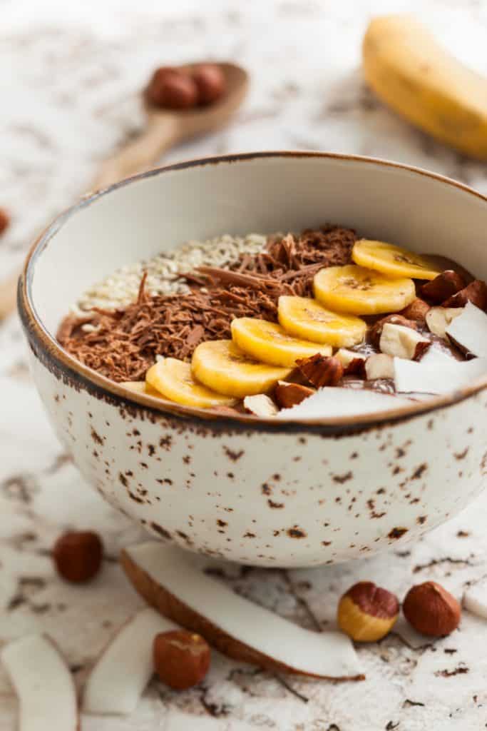 Chocolate hazelnut smoothie bowl topped with sliced banana, shredded coconut, chopped  chocolate, nuts and sesame seeds. Soft focus
