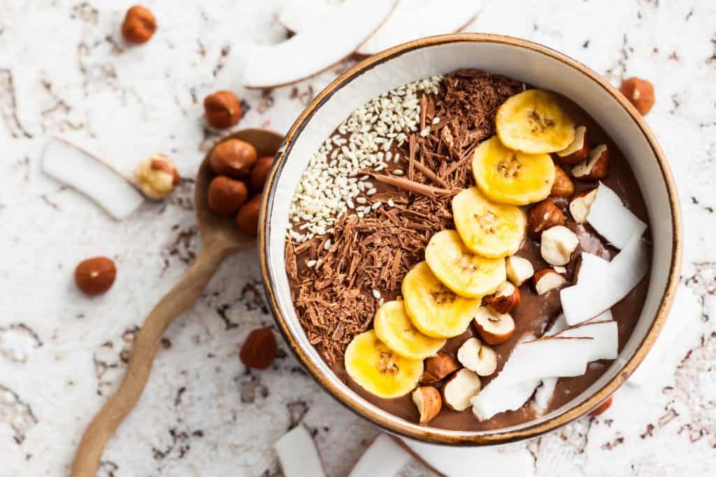 Chocolate hazelnut smoothie bowl topped with sliced banana, shredded coconut, chopped  chocolate, nuts and sesame seeds.