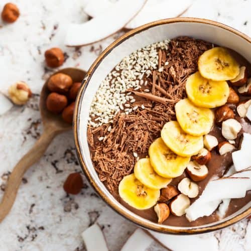 Chocolate hazelnut smoothie bowl topped with sliced banana, shredded coconut, chopped chocolate, nuts and sesame seeds.