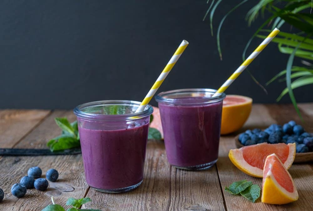 two blueberry detox smoothies on wooden background with ingredients surrounding