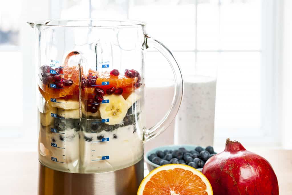 Prepared smoothies and healthy smoothie ingredients in blender with fresh fruit ready to blend on kitchen table