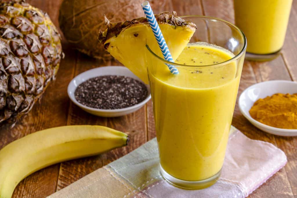 Close up fresh blended fruit smoothies surrounded by raw ingredients in drinking glass with pineapple slice garnish and blue striped straw
