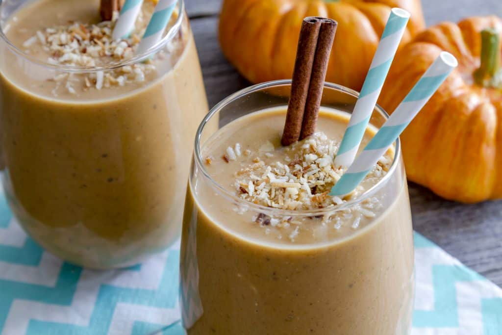 Close up of two fresh pumpkin coconut smoothies garnished with toasted coconut sitting on blue chevon napkin and blue swirled straws and cinnamon sticks