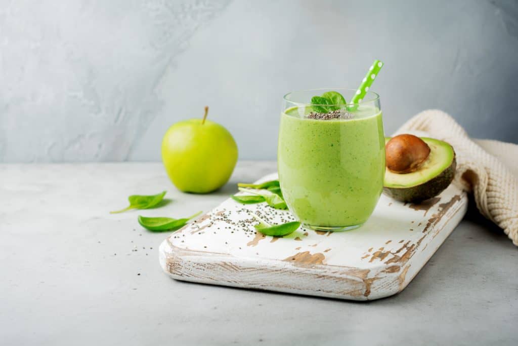 apple avocado smoothie in glass on wooden board with apple, avocado and spinach around