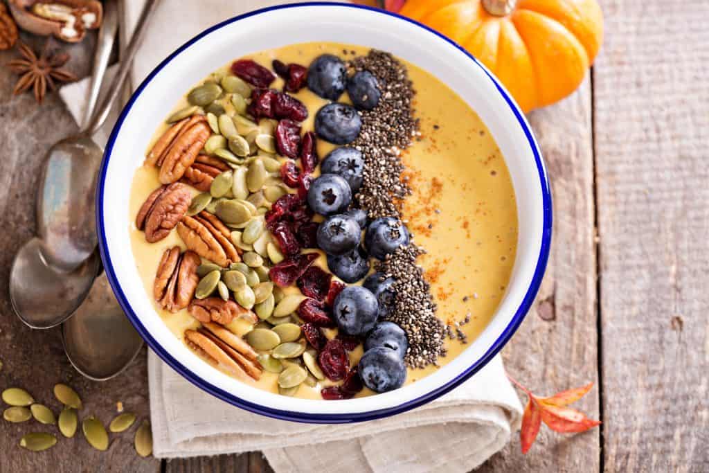 pumpkin smoothie bowl on table with toppings of fresh and dried fruit, seeds and nuts