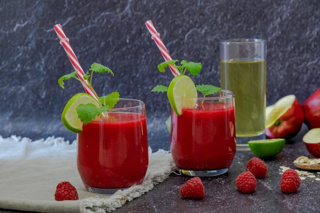 two apple raspberry smoothies in glasses with ingredients in background