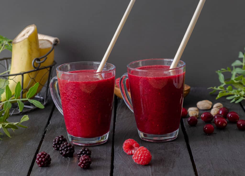 two blackberry raspberry smoothies in glasses with straws and ingredients surrounding