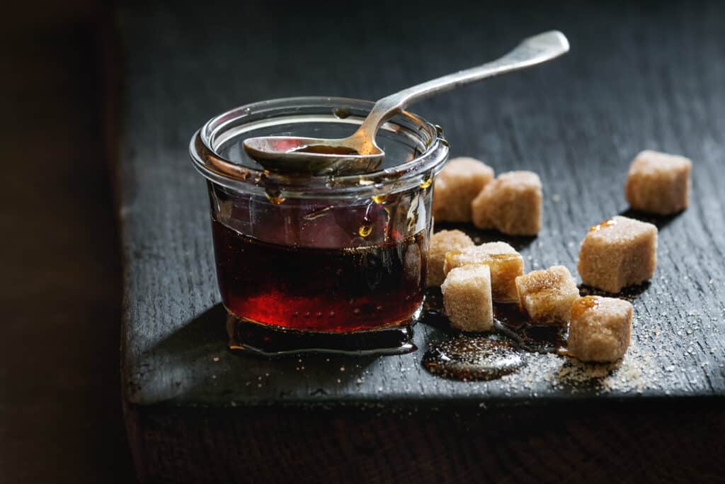 Homemade brown sugar syrup in glass jar standing on black wooden board with spoon and can sugar cubes.