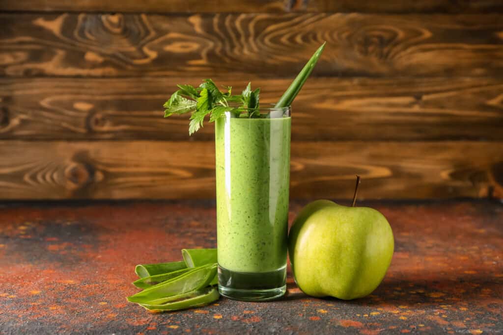 glass of aloe vera smoothie in front of wooden background with aloe and apple