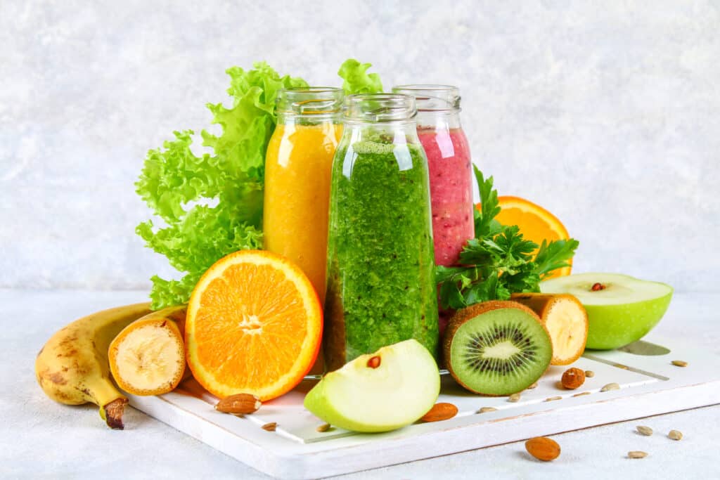 Green, yellow, purple smoothies in currant bottles, parsley, apple, kiwi, orange on a gray table